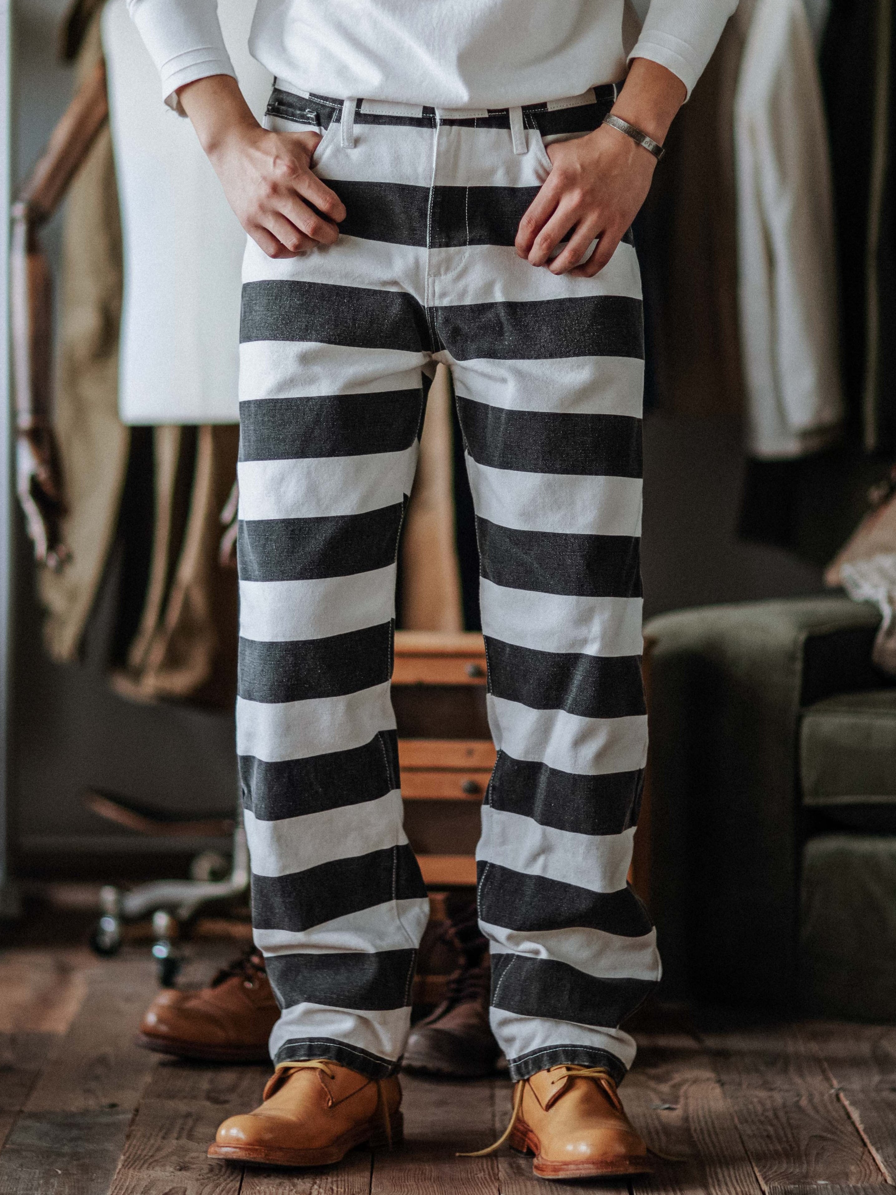A person standing on a wooden floor wearing wide striped prison pants with a black and white colorway, paired with a white shirt and brown shoes.